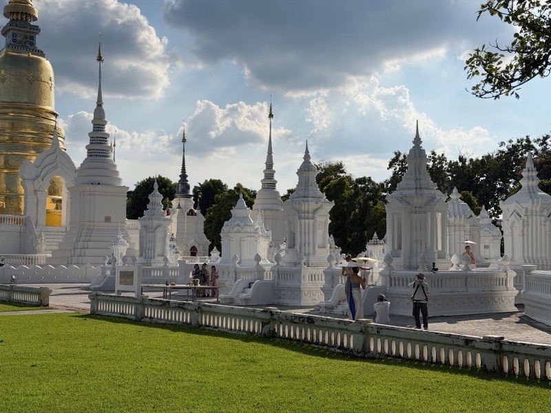 The white spired ku (reliquary stupas) of the northern royal family at Wat Suan Dok, Chiang Mai, beside a large golden chedi, under a cloudy sky with a green lawn in front