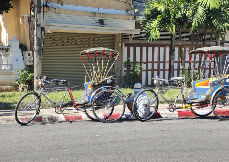 Two pedal samlors parked side by side on a street near Kad Luang market in Chiang Mai, with canvas roofs and brightly coloured seats