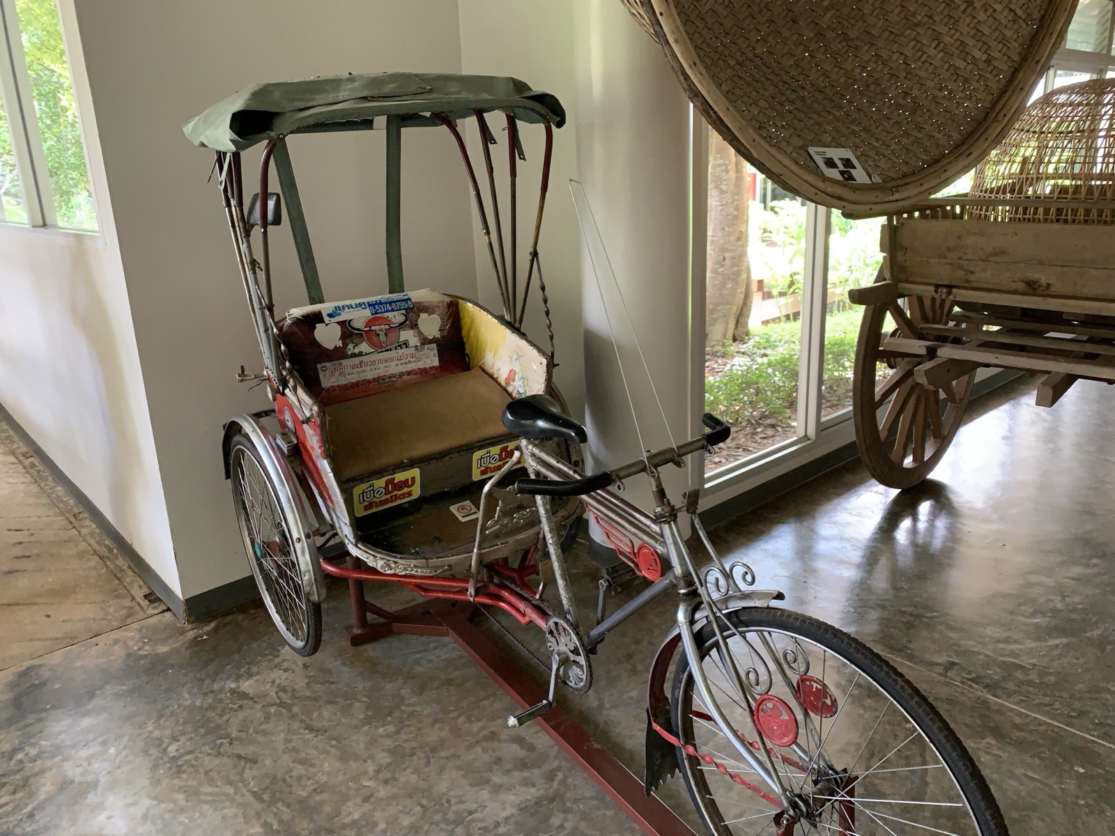 An old pedal samlor on display in a museum, with a green canvas roof, a brown seat and stickers on the body