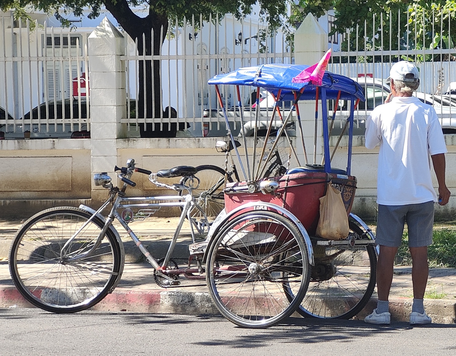 A pedal samlor parked beside a bicycle on the pavement near Kad Luang, Chiang Mai, with a blue canvas roof and a pink flag