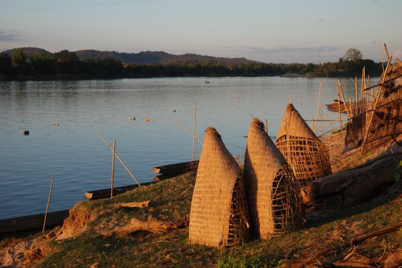 Large cone-shaped woven bamboo fish traps lined up on a river bank in Laos, with a small wooden boat and distant hills in the evening light