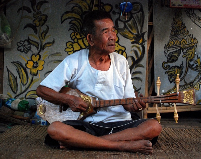 ครูดนตรีพื้นเมืองนั่งขัดสมาธิบนเสื่อ ดีดซึงไม้แกะลายสีทอง ที่บ้านในอำเภอแม่ออน เชียงใหม่