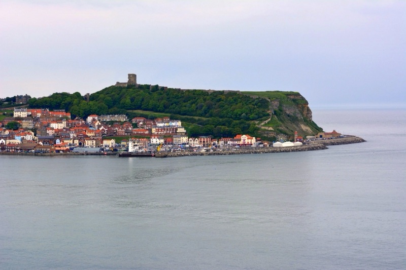 The town of Scarborough in northern England, with orange-roofed houses along the bay and a ruined castle on the green headland above a calm sea