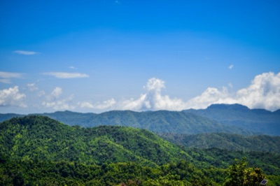 A ridge of green forested hills under a bright blue sky, with white clouds piled above the peaks