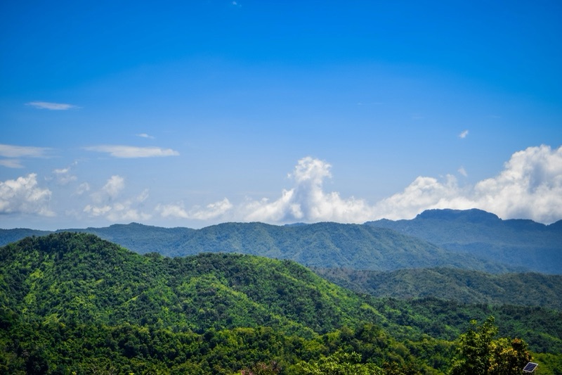 A ridge of green forested hills under a bright blue sky, with white clouds piled above the peaks