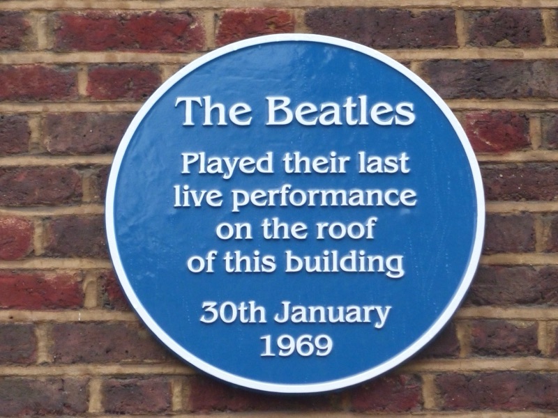 ป้ายวงกลมสีน้ำเงินติดกำแพงอิฐ มีข้อความภาษาอังกฤษว่า The Beatles played their last live performance on the roof of this building, 30th January 1969