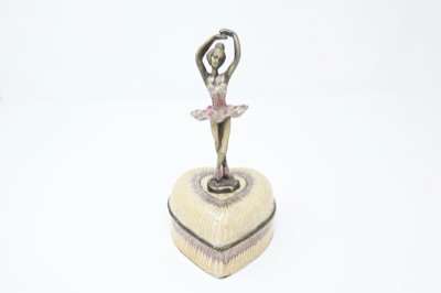 An ivory heart-shaped music box with a small ballerina figure standing on its lid, on a white surface