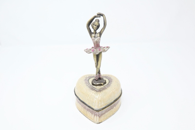 An ivory heart-shaped music box with a small ballerina figure standing on its lid, on a white surface