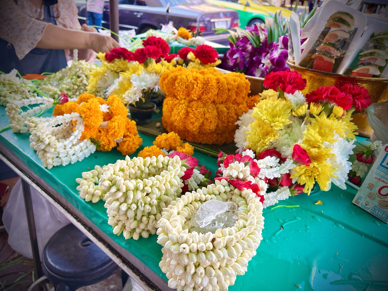 พวงมาลัยดอกมะลิสีขาวหลายพวงวางบนโต๊ะสีเขียว ข้างพวงมาลัยดาวเรืองสีส้มและช่อดอกไม้สีแดงเหลือง