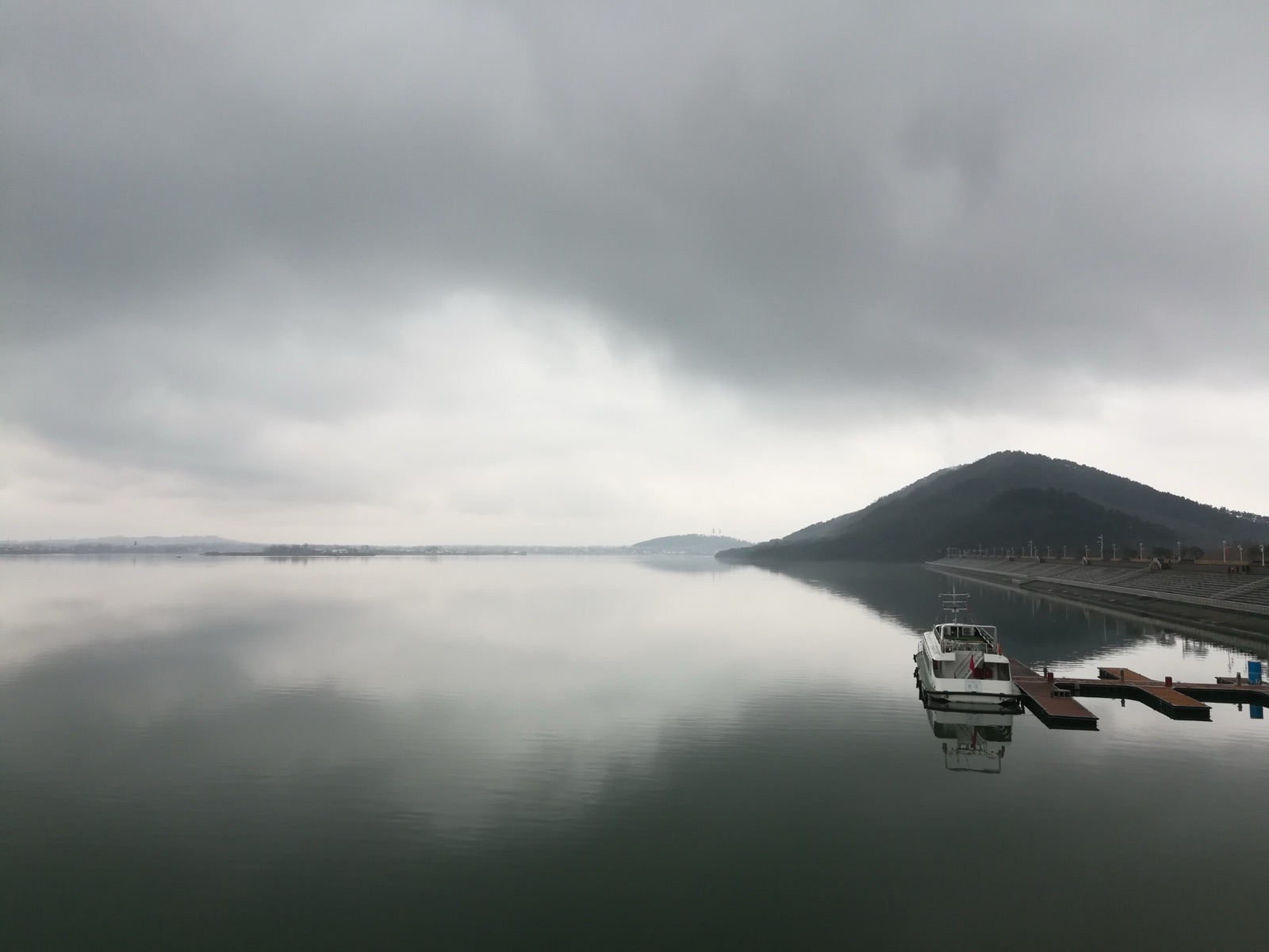 ทะเลสาบกว้างนิ่งใต้ท้องฟ้าครึ้ม มีเรือเล็กจอดที่ท่าและเนินเขาอยู่ริมน้ำ
