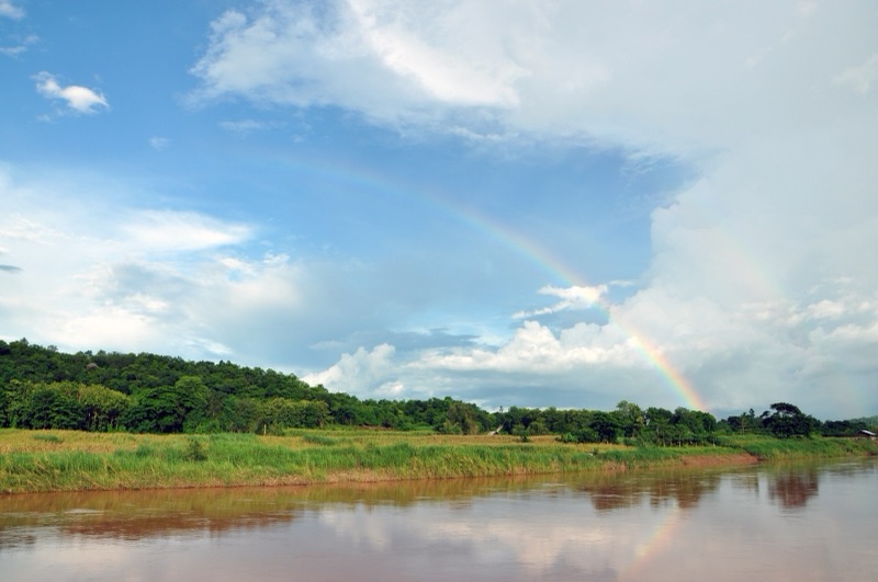 A rainbow arching across a blue sky with white clouds over the Wa River, grassland and green forest in Nan province