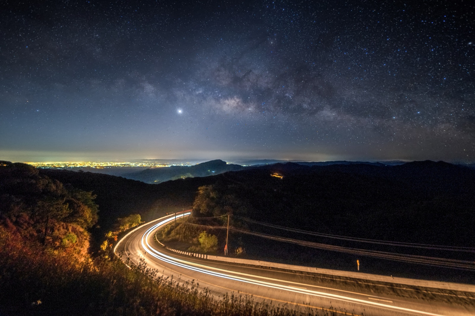 A night sky full of stars and the Milky Way above a winding mountain road traced by car light trails, with distant city lights