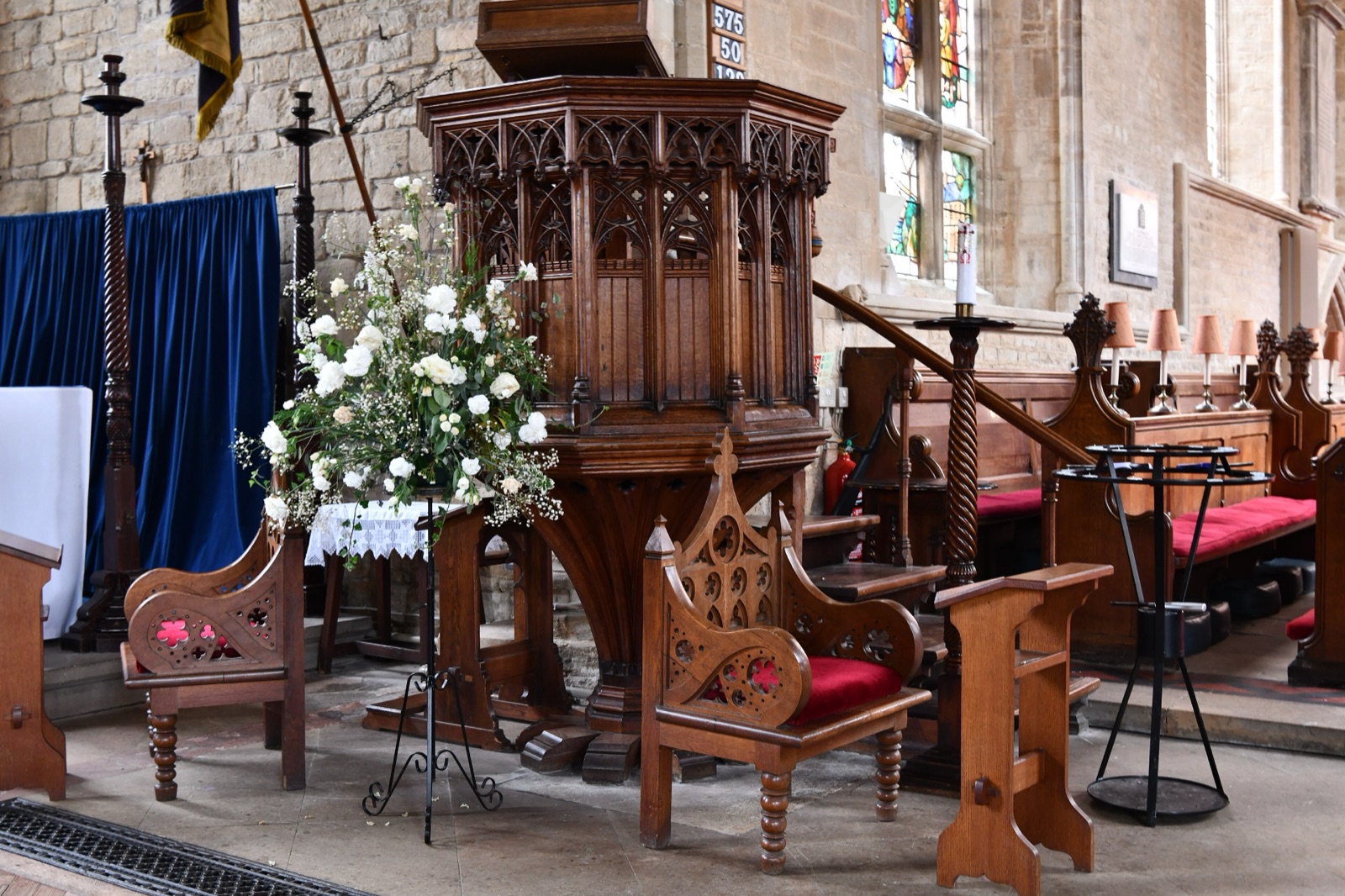 A carved octagonal wooden pulpit in Olney church, with a vase of white flowers and wooden chairs with red cushions in front