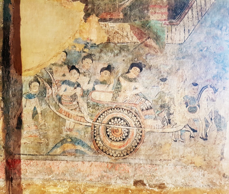 An old mural at Wat Phumin in Nan province showing men and women riding in a cart with flower-patterned wheels, with a walking figure and a horse beside it