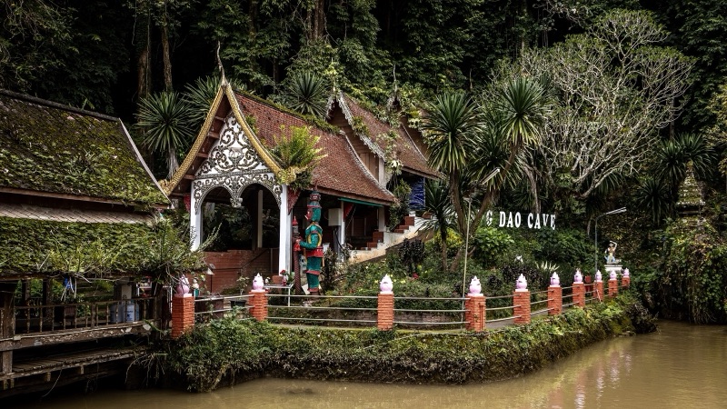 The way up to Wat Tham Chiang Dao: a Lanna-style pavilion among dense green trees beside a pond, with a row of orange fence posts
