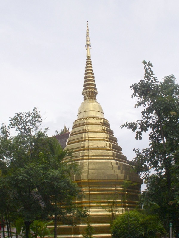 A tall golden bell-shaped chedi with a slender spire among green trees under an overcast sky