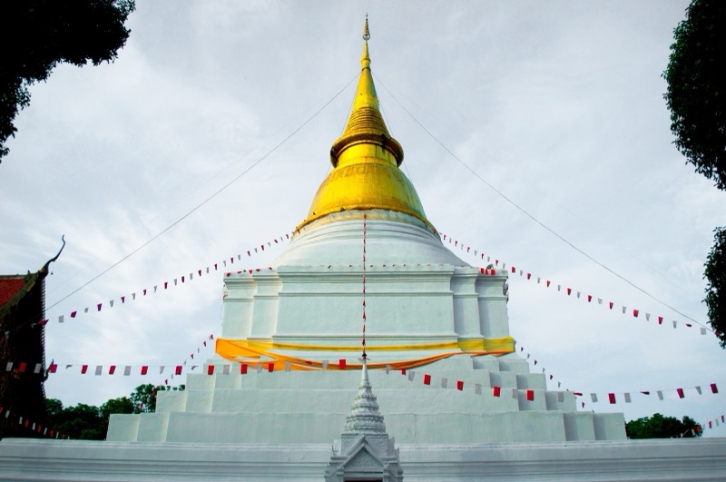 A chedi with a white base and golden spire, wrapped in yellow cloth, with red and white bunting strung from the top