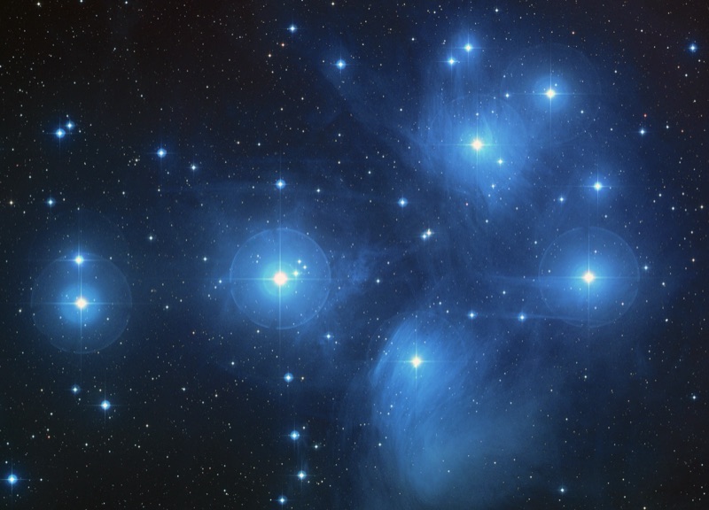 The Pleiades star cluster: several bright blue stars grouped together, wrapped in faint blue nebula on a dark sky