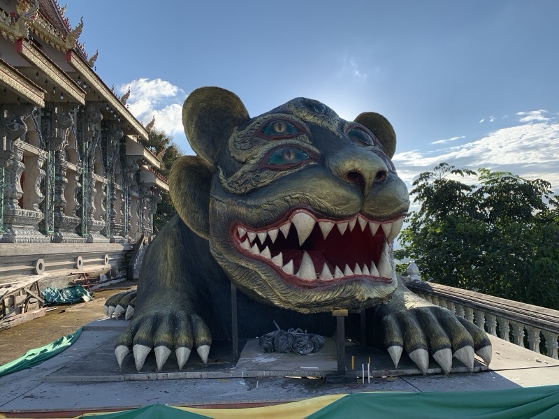 A large dark-green statue of Maeng Si Hu Ha Ta with a tiger-like face and bared fangs, crouching beside a hall at Wat Phra That Doi Khao Khwai Kaeo, Chiang Rai
