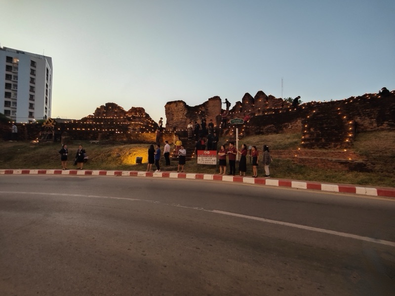 แถวประทีปเรียงเป็นเส้นเรืองแสงบนกำแพงเมืองเชียงใหม่ยามพลบค่ำในคืนก่อนวันแรกของยี่เป็ง มีผู้คนยืนอยู่ไกล ๆ ริมถนน