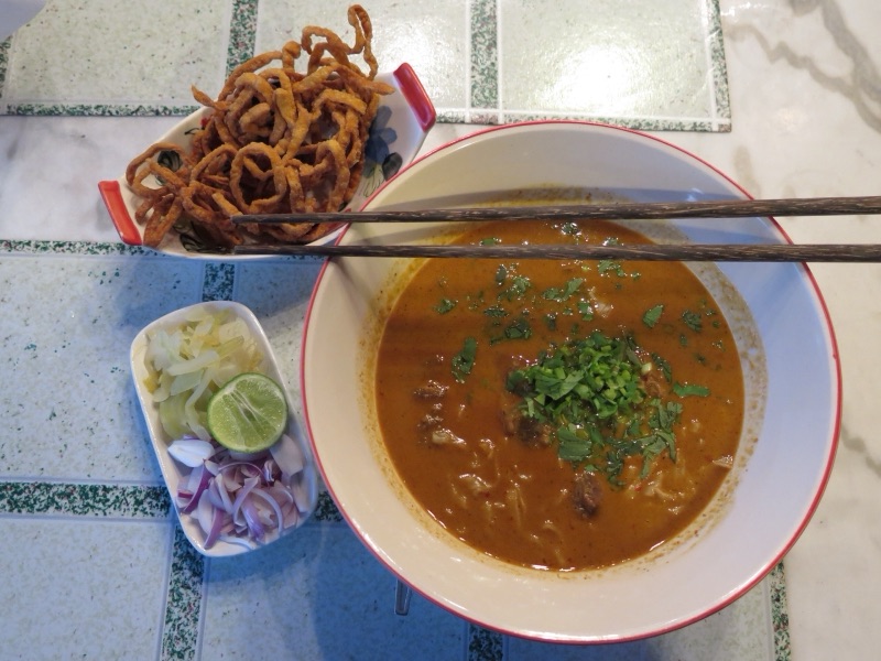 A bowl of khao soi with crispy noodles, pickled mustard greens, shallots and lime on the side