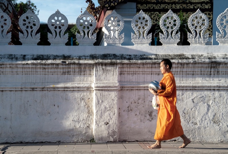 A monk in an orange robe carries his alms bowl past a white temple wall