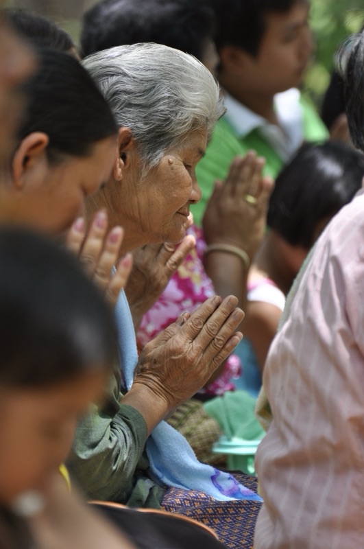 A white-haired elderly woman makes a wai among a crowd of people with their palms joined