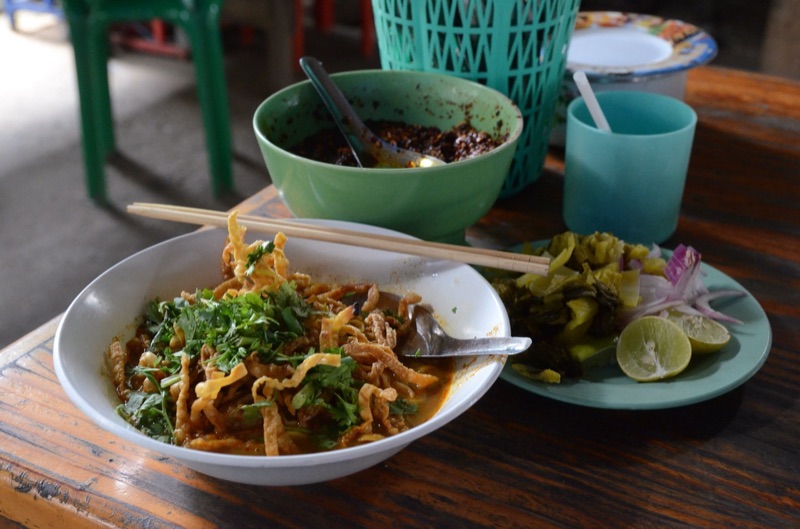 ข้าวซอยในชามขาว โรยหมี่กรอบและผักชี วางคู่จานผักกาดดอง หอมแดง และมะนาว บนโต๊ะไม้ของร้านข้าวซอยในเชียงใหม่