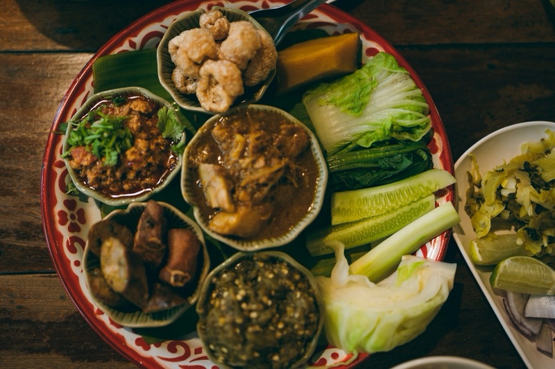 สำรับอาหารเหนือในถาดลายคราม มีแคบหมู ไส้อั่ว น้ำพริกหนุ่ม แกงในกระทงใบตอง และผักสดวางรวมกันบนโต๊ะไม้