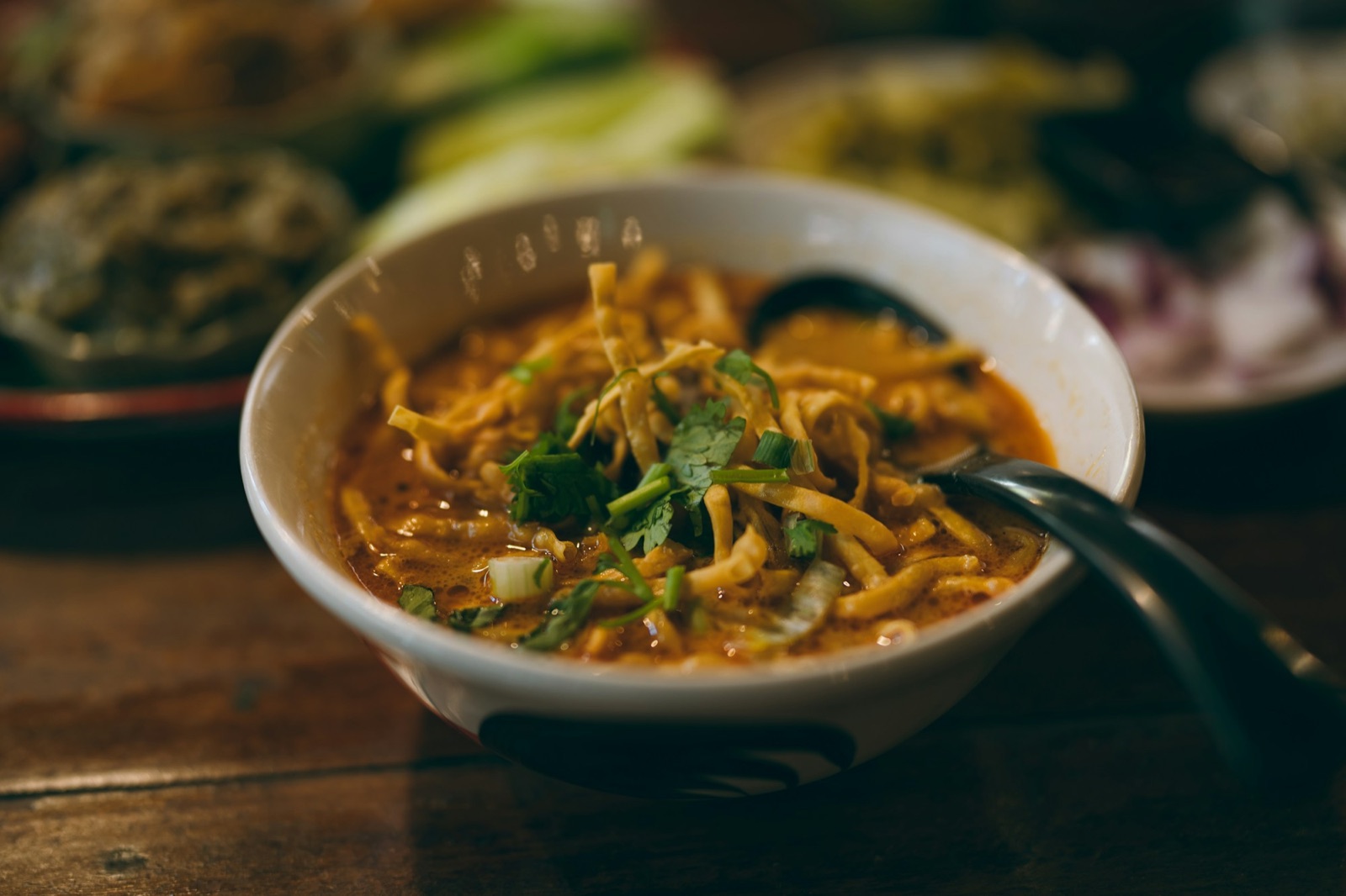ข้าวซอยในชามสีขาว โรยผักชีและเส้นกรอบ มีช้อนสีดำวางพาดในชาม