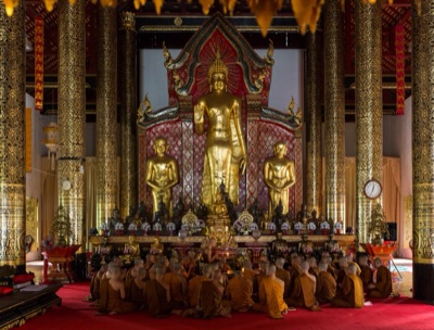 พระสงฆ์นั่งสวดมนต์เรียงแถวในวิหาร หน้าพระพุทธรูปยืนสีทอง