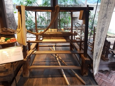 A traditional wooden floor loom with cloth in progress and treadles below