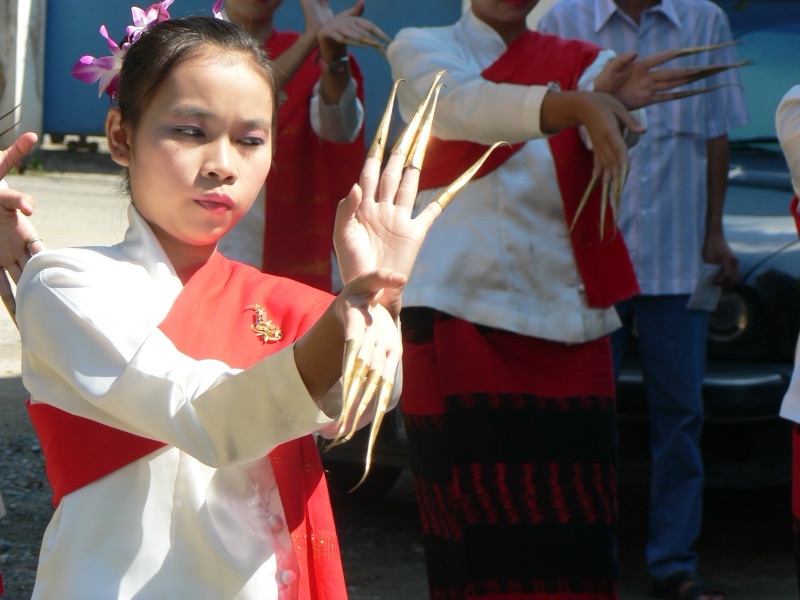 A young dancer in a white blouse and red sash, wearing long nail extensions on every finger, raising her hands to face level during a fingernail dance in Chiang Mai