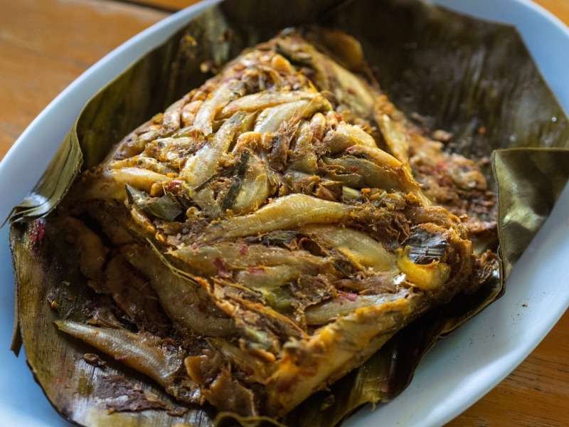 Aep pla noi unwrapped from its banana leaf on a white plate, with small fish lying in the curry paste
