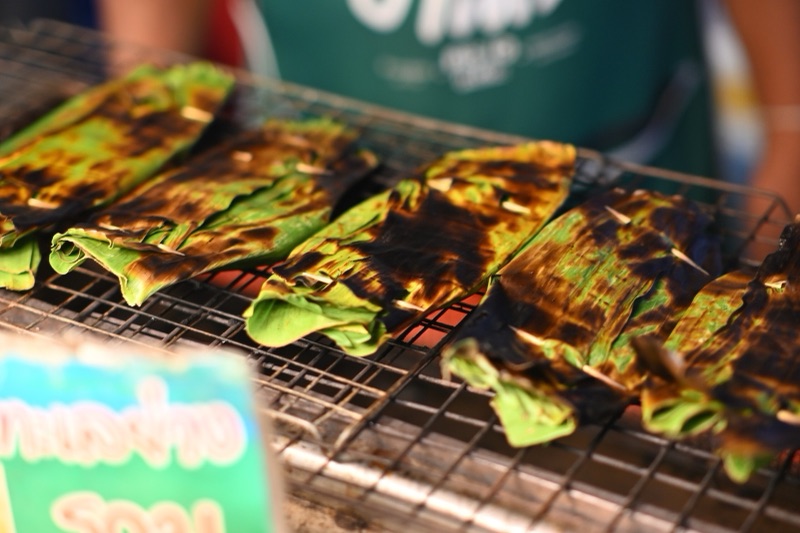 Snacks wrapped in banana leaf grilled on a metal rack, the leaves charred black in places