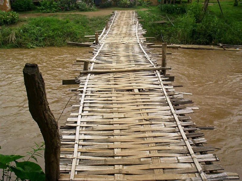 A bridge deck of woven bamboo panels across a muddy brown river, with a wooden post on the left